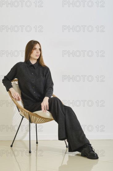 A woman sits gracefully in a wicker chair, wearing a trendy black cardigan and loose pants. The practical style highlights her ease and confidence in a minimalistic indoor space