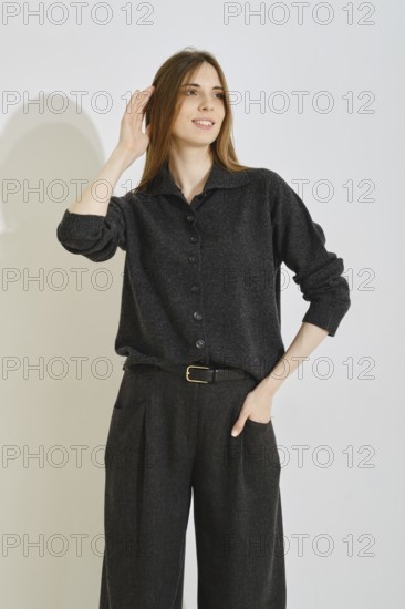 A young woman playfully touch her smooth long hair wearing trendy outfit featuring a dark button-up shirt and loose trousers. The setting is bright and minimalist, emphasizing style and comfort