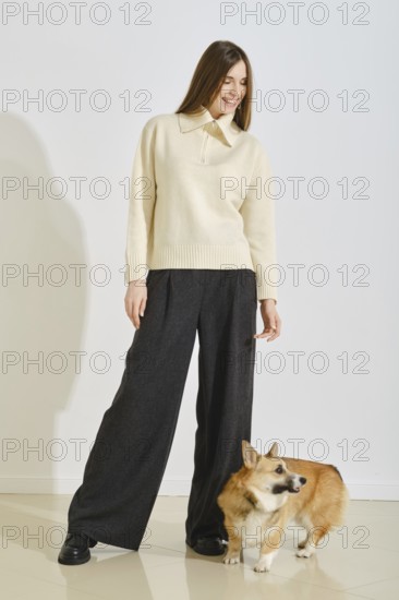 Playful woman in ivory sweater and loose pants playing with her corgi dog in bright room