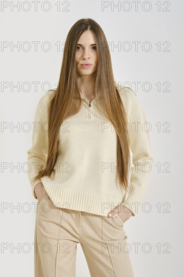 A woman in a cream sweater poses with her hands in pockets of beige pants. Her long hair flows freely as she stands against a light wall, embodying style with ease and comfort during daytime activities