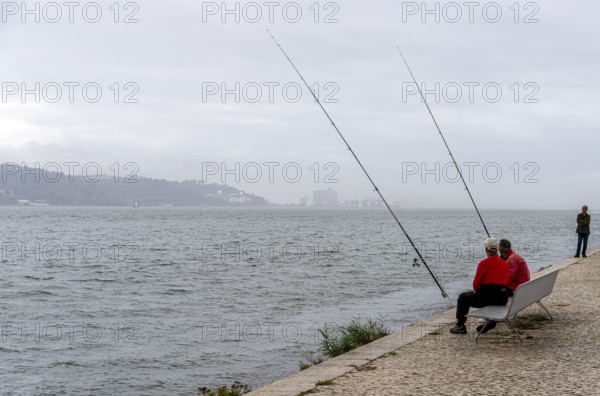 Anglers on Ponte 25 de Abril, 3.2 km long bridge in Portugal with a 2278 meter long suspension bridge across the Tagus, cloudy and rainy weather, third-longest suspension bridge with combined road and rail traffic, Lisbon, Portugal