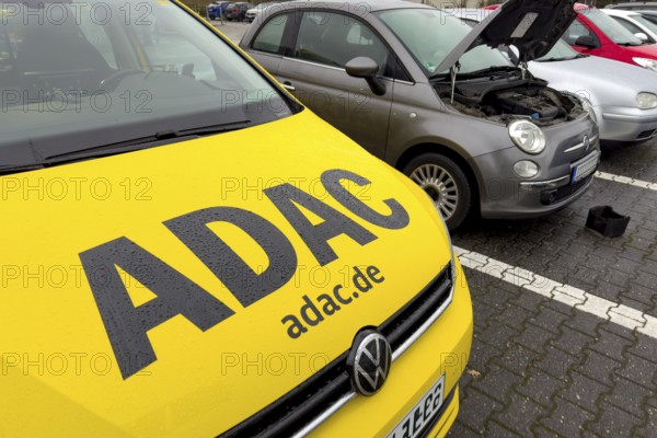 Front car with large ADAC roadside assistance lettering on vehicle for roadside assistance, behind car with open bonnet open in parking lot, replacement battery packaging in front of it, Germany