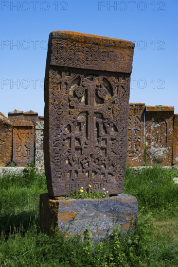 A detailed Armenian crossstone with engravings in front of several others, surrounded by grass under a clear sky, crossstones, khachkar, world's largest khachkar field, Noratus cemetery, Noradus, Gegharkunik province, Armenia