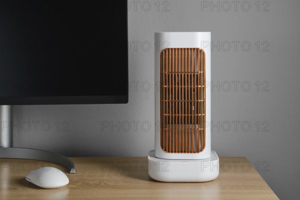 A modern electric heater is positioned on a wooden desk next to a computer monitor. The warm glow of the heater adds comfort to the indoor workspace, perfect for cold weather
