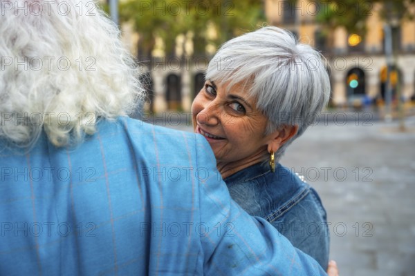 Senior woman with short gray hair smiling over her partner's shoulder as they embrace on a lively city street, sharing a joyful, affectionate moment outdoors