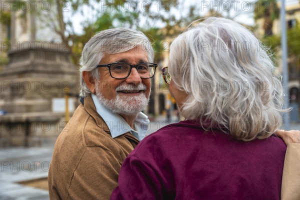 Senior man with a beard and glasses smiling warmly at his female partner, enjoying time together outdoors in an urban setting, representing a concept of lasting love and companionship in retirement