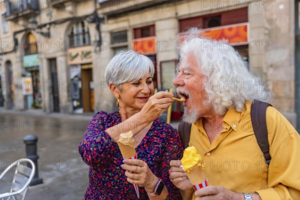 Senior couple on a charming european street sharing gelato, woman feeding man a cone as they stroll, smiling and enjoying a sweet, romantic moment during vacation