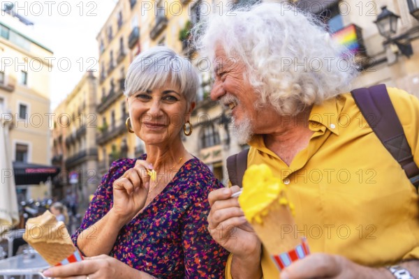 Happy mature couple smiling together while eating ice cream cones, experiencing leisure and love during a city break, with a pride flag representing inclusivity visible in the background