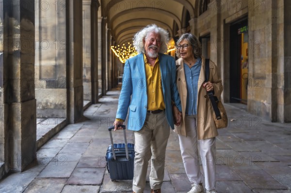 Happy senior couple smiling and holding hands while walking through a european city street, pulling luggage and enjoying an active retirement vacation together exploring architecture and sights