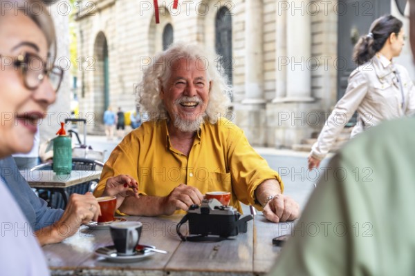 Mature man with white hair and beard laughing brightly while enjoying coffee and conversation with friends at an outdoor cafe, sharing moments of joy and connection in an urban setting
