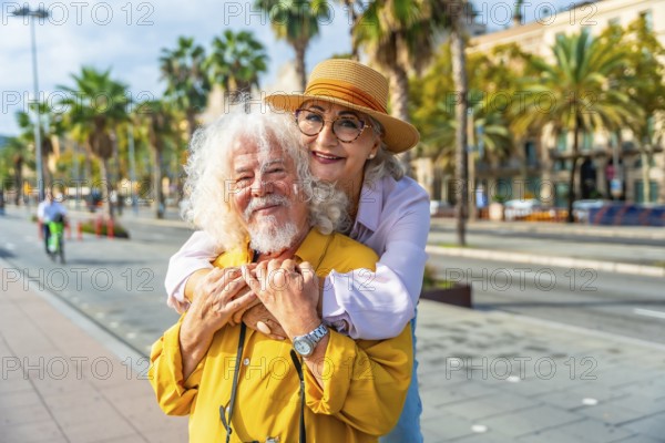 Senior couple embracing and smiling at the camera on a sunny city promenade with palm trees and buildings, enjoying vacation, retirement, and happy, loving companionship