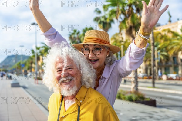Senior couple enjoying their summer vacation together in a vibrant city, smiling brightly and raising arms in a gesture of joy and freedom while traveling