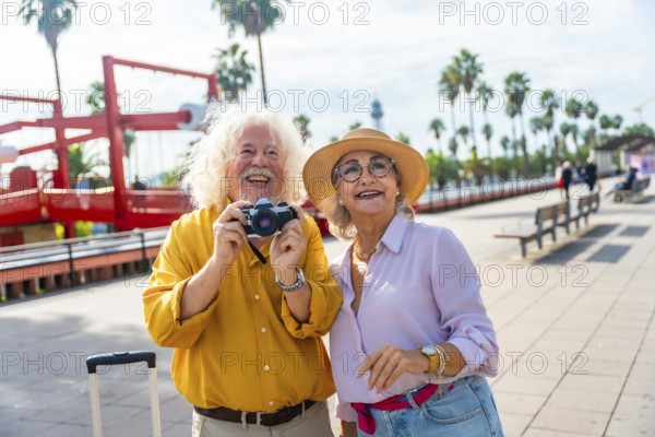 Happy senior couple enjoying their vacation and exploring a sunny city while traveling, capturing memories with a vintage camera on a beautiful boardwalk