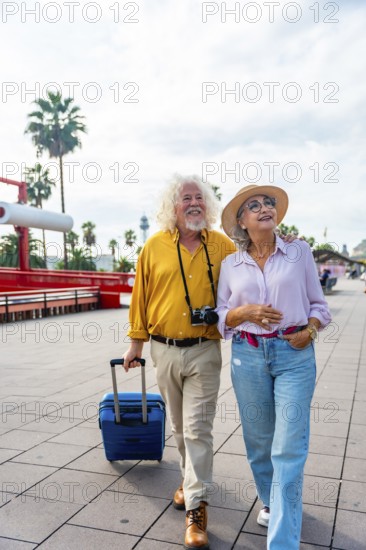 Senior couple walking on a city promenade, smiling and looking up, carrying a suitcase and a camera, enjoying their vacation and exploring new places together
