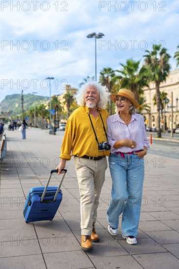 Smiling senior couple embracing a happy retirement, walking together with their luggage through a sunny city street, exploring and enjoying an exciting travel adventure
