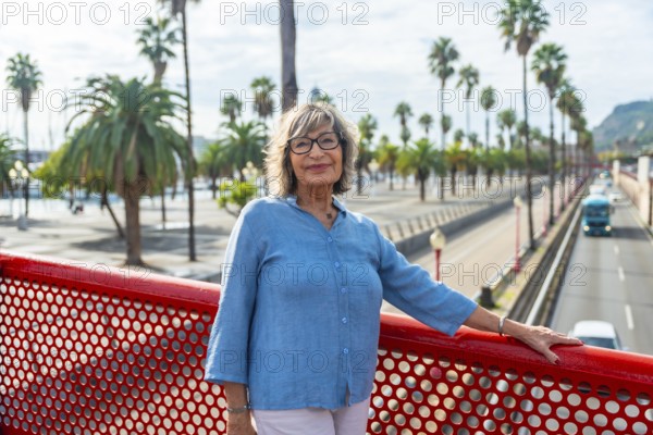 Senior woman standing on a red bridge, smiling directly at the camera, with a vibrant city street and numerous palm trees forming the blurred background