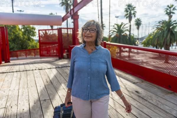 Senior woman walking across a pedestrian bridge with her luggage, enjoying an urban vacation on a sunny day with palm trees and a marina in the background