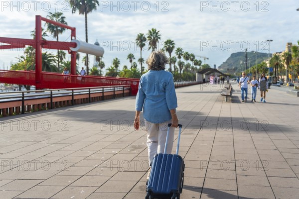 Senior woman walking with a suitcase along a sunny boardwalk lined with palm trees and a distinctive red bridge, enjoying a relaxed vacation or leisure trip