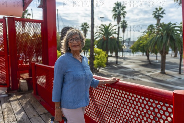 Senior woman standing on red bridge at a sunny harbor, smiling confidently in glasses while enjoying leisure travel and sightseeing among palm trees and boats in barcelona port