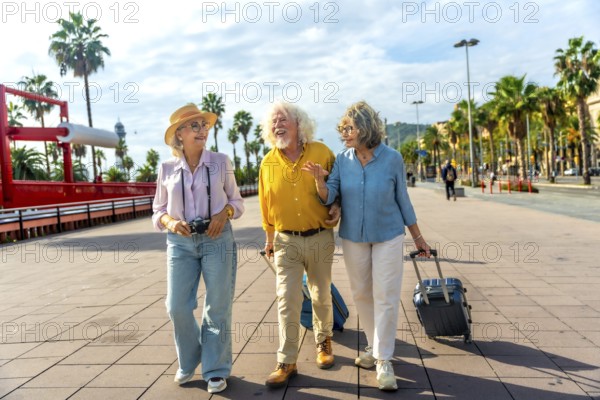 Three cheerful senior tourists walking together on a promenade with palm trees, carrying luggage and a camera, enjoying friendship and travel during their holiday