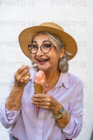 Happy senior woman wearing a straw hat and stylish glasses, smiling while eating a refreshing ice cream cone with a spoon during a pleasant summer day
