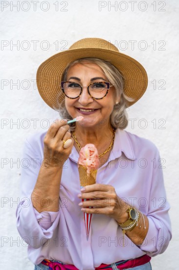 Happy senior woman wearing a straw hat and glasses smiling while eating a refreshing ice cream cone with a small spoon, celebrating moments of joy and relaxation in summer
