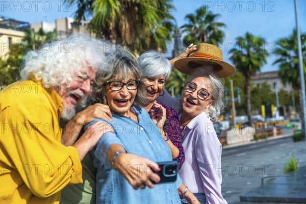 Group of four joyful senior friends laughing and smiling while capturing a memorable selfie with a smartphone outdoors, enjoying their friendship and freedom during a sunny day in the city