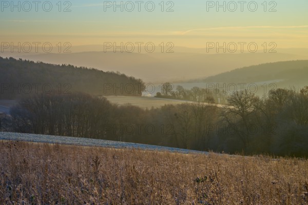 Frozen meadow in morning light with a foggy valley and rolling hills in the background, Seckmauern, Lützelbach, Maintal, Odenwald, Hesse, Germany
