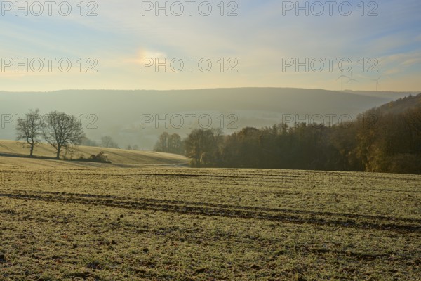 View over a frosty field and distant hills with wind turbines on the horizon at dawn, Seckmauern, Lützelbach, Odenwald, Hesse, Germany