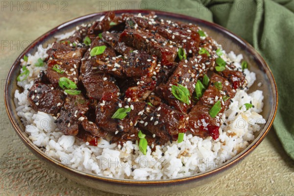 Crispy beef, with green onions and sesame seeds, on a rice pad, in a bowl with chopsticks, on a textured surface, no people