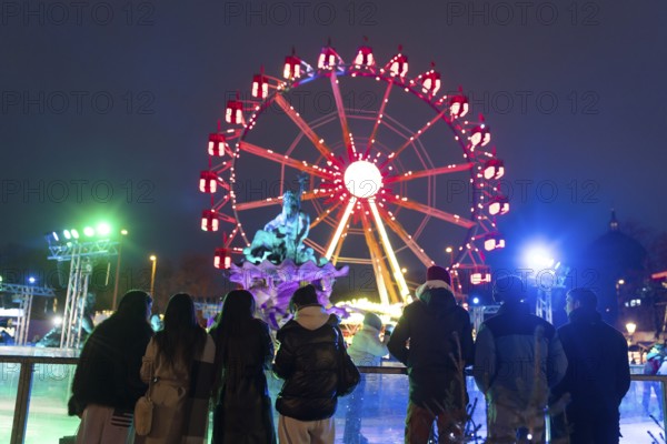 Several people watch ice skaters in front of the Neptune Fountain and a Ferris wheel at the Berlin Christmas market on Alexanderplatz, Berlin, 24.11.2025