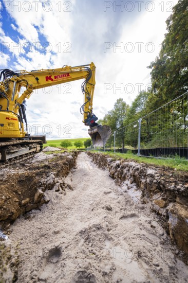 A yellow excavator digs a deep ditch surrounded by nature and a cloudy sky, Bau PV Freifaechenanlage, Weil der Stadt, Germany