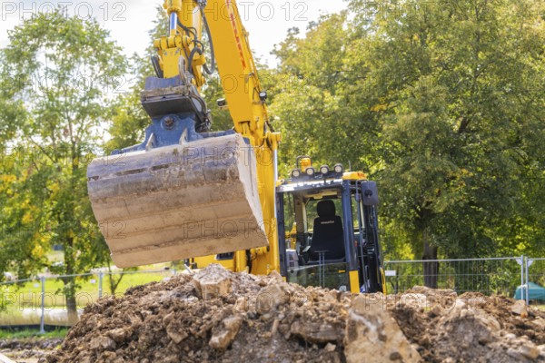 An excavator on a construction site lifts soil near trees and a cloudy sky, building a PV open air system, Weil der Stadt, Germany