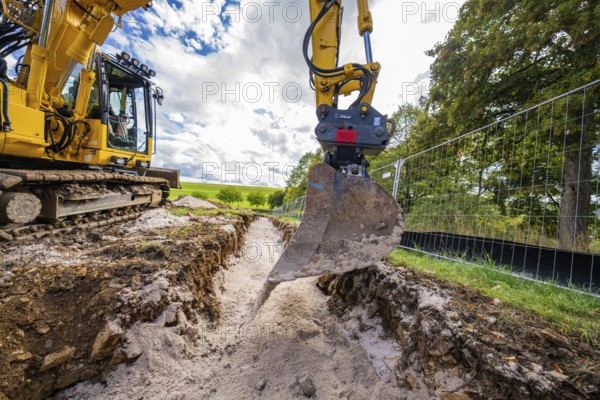 An excavator lifts soil from a deep ditch on a construction site in cloudy skies, construction PV Freifaechenanlage, Weil der Stadt, Germany