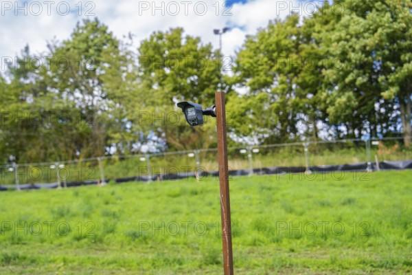 Camera on a post with grass and trees in the background under a blue sky, Bau PV Freifaechenanlage, Weil der Stadt, Germany