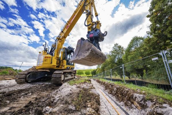 An excavator excavates a ditch in a natural environment under cloudy skies, Bau PV Freifaechenanlage, Weil der Stadt, Germany