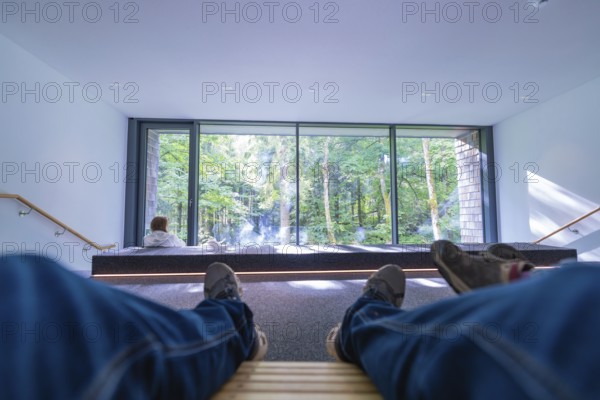 Person relaxing on wooden loungers in a modern room with a large glass front and a view of the forest, Ruhestein National Park Center, Black Forest, Baiersbronn, Germany