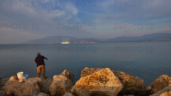 A fisherman fishing on rocky shore with a large sailing ship in the background in grey weather, Nauplion, Nafplion, Peloponnese, Greece