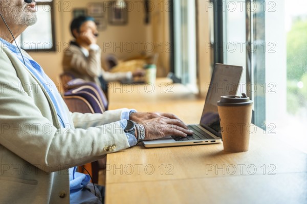 Senior professional working remotely at a busy coworking table, focused on laptop and video call with distant colleague, headset on, coffee nearby, modern flexible workspace