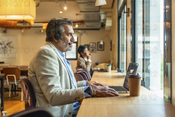 Mature man wearing a headset and working on a laptop in a modern coworking space, focused on a video call or customer service chat while telecommuting remotely