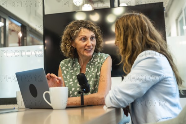 Two professional women collaborating on a podcast, recording a conversation with a microphone and laptop in a contemporary coworking space, focusing on content creation and communication