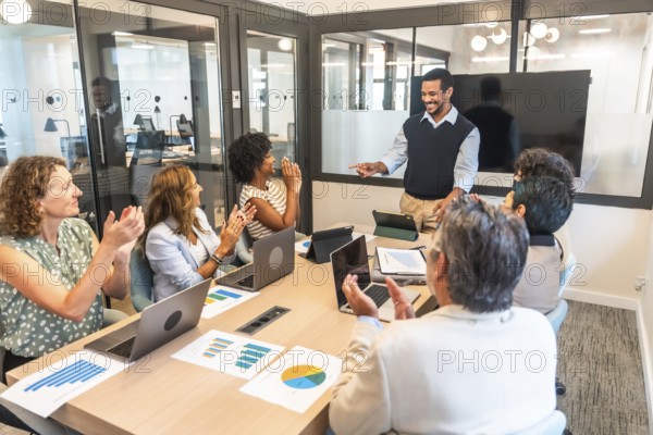 Diverse business team in a modern boardroom applauding a smiling presenter, celebrating project success and teamwork during a collaborative meeting with laptops and charts on the table