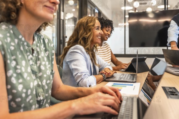 Diverse group of smiling businesswomen collaborating and engaging in productive work during a team meeting, using laptops and reviewing data in a contemporary coworking office environment