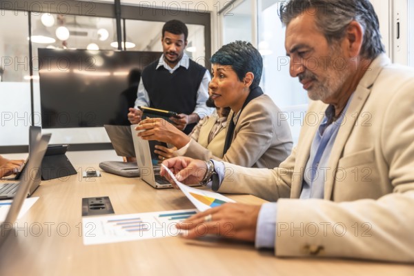 Business colleagues of diverse ages and ethnicities collaborating during an important meeting, reviewing financial data on laptops and printed charts in a modern corporate coworking space