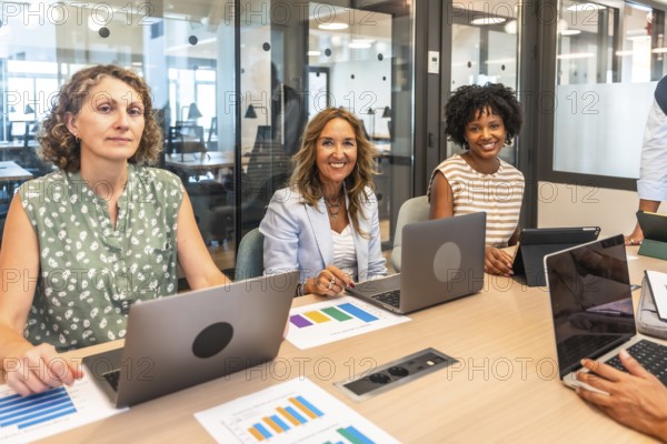 Diverse businesswomen are smiling at the camera, working with laptops and analyzing data graphs while collaborating around a table in a modern office meeting room