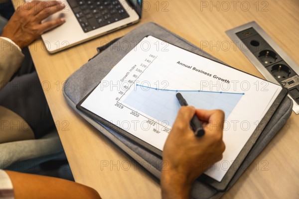 Business colleagues collaborating on strategy, analyzing a printout annual business growth chart and financial data during a meeting, with one person pointing to the graph and another using a laptop