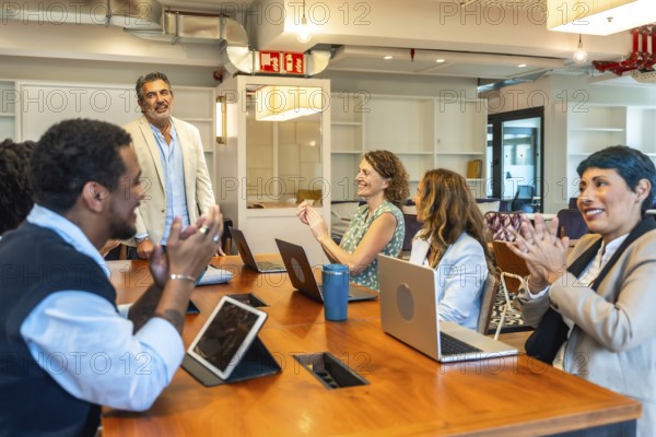 Diverse business colleagues sitting around a large wooden table, smiling and clapping for a speaker after a successful presentation in a modern coworking office space
