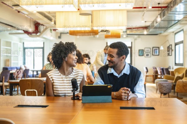 Two diverse colleagues collaborating and discussing ideas while recording a podcast with a microphone and digital tablet in a modern coworking office space during a business meeting