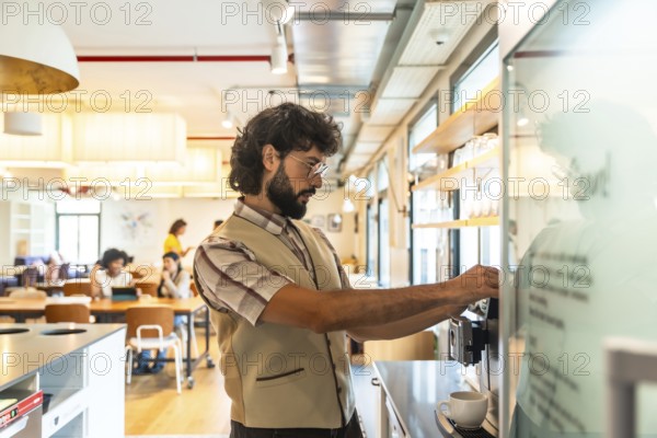 Bearded man preparing coffee at a modern coworking space, taking a relaxed break from work while colleagues chat in the bright, stylish shared office background