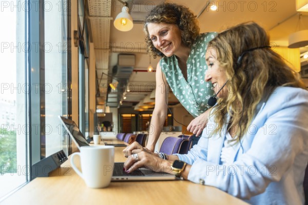 Two smiling women are collaborating and supporting each other while wearing headsets, working on laptops at a coworking office, demonstrating teamwork and professional synergy